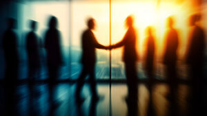 Silhouettes of people in a hallway with two central figures shaking hands as a bright light glows behind them, suggesting a business agreement.