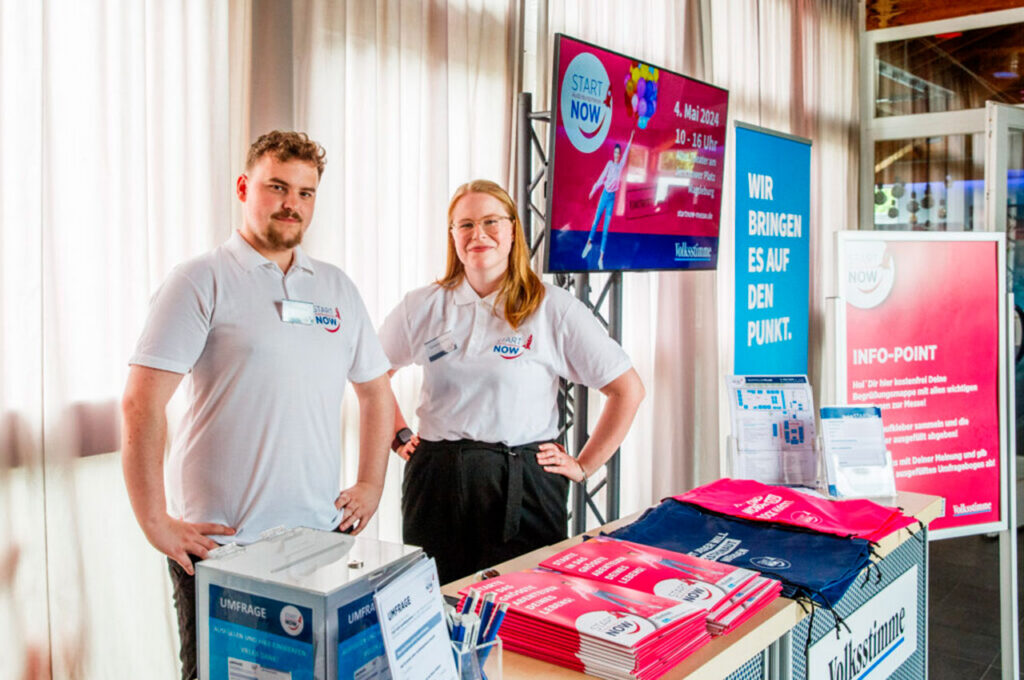 Informationsstand auf einer Karrieremesse mit zwei Personen hinter einem Messestand. Auf dem Tresen liegen Flyer und Werbematerialien, im Hintergrund sind Informationsdisplays und Roll-ups zu sehen. ©Mediengruppe Mitteldeutschland